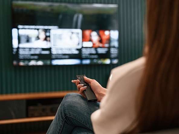 woman holds remote control pointing at TV, displaying a blurred selection of channels